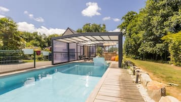 Une piscine entourée de parois en verre, dotée d’une terrasse en bois et bordée de verdure.