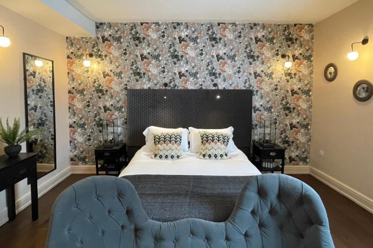 Quarto com cama grande, duas mesas de cabeceira, parede com padrão floral e uma poltrona azul.