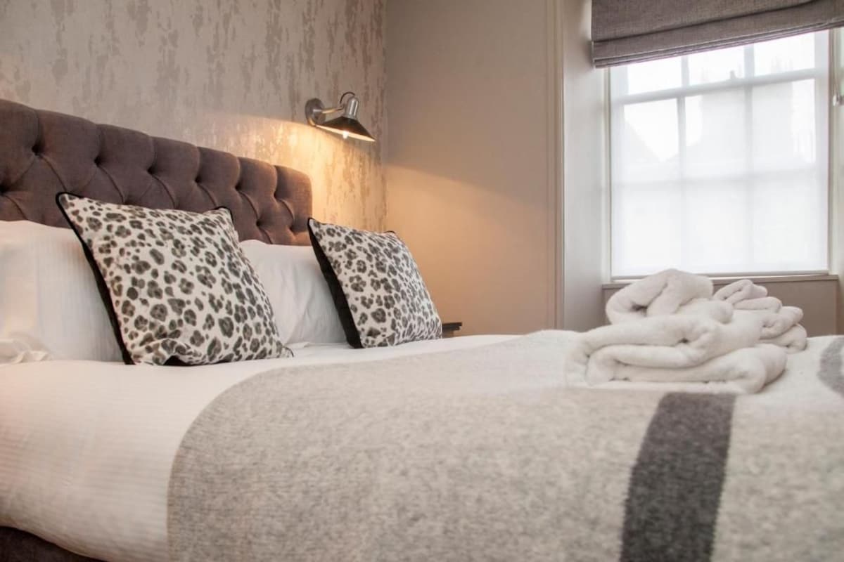 Quarto com cabeceira estofada, duas almofadas com padrão de leopardo, colcha branca e uma pilha de toalhas brancas.