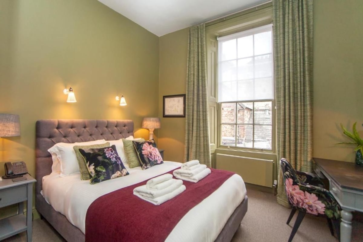 Quarto de hotel com uma cama grande, duas luminárias de cabeceira, uma janela com cortinas, uma mesa de trabalho e uma cadeira.