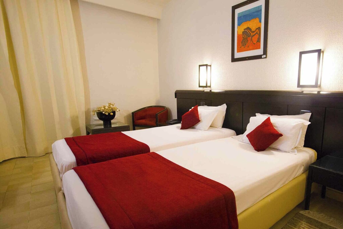Une chambre d’hôtel avec deux lits, une table de chevet, une chaise, un vase avec des fleurs et un tableau au mur.