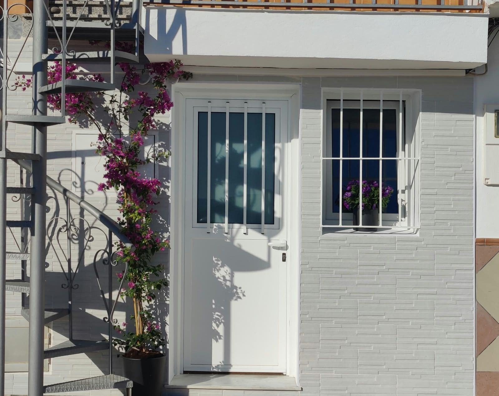Un edificio blanco con una puerta y una ventana, un balcón con barandillas de metal y una planta en maceta con flores moradas.