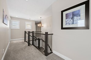 A hallway with a carpeted floor, a black railing, and a framed picture on the wall. - 3BR Main & Upper Floor in Airdrie | Sleeps 9 | Canal View (Airdrie)