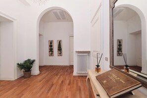 Passadís amb terra de fusta, parets blanques i un gran mirall. Hi ha dues plantes en testos, un radiador i un llibre sobre una taula.
