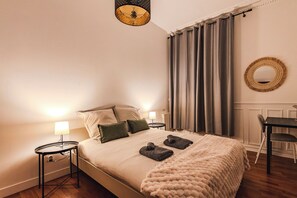 Um quarto bem organizado com uma cama, duas mesas de cabeceira, uma cadeira, um espelho redondo e uma janela com cortinas.