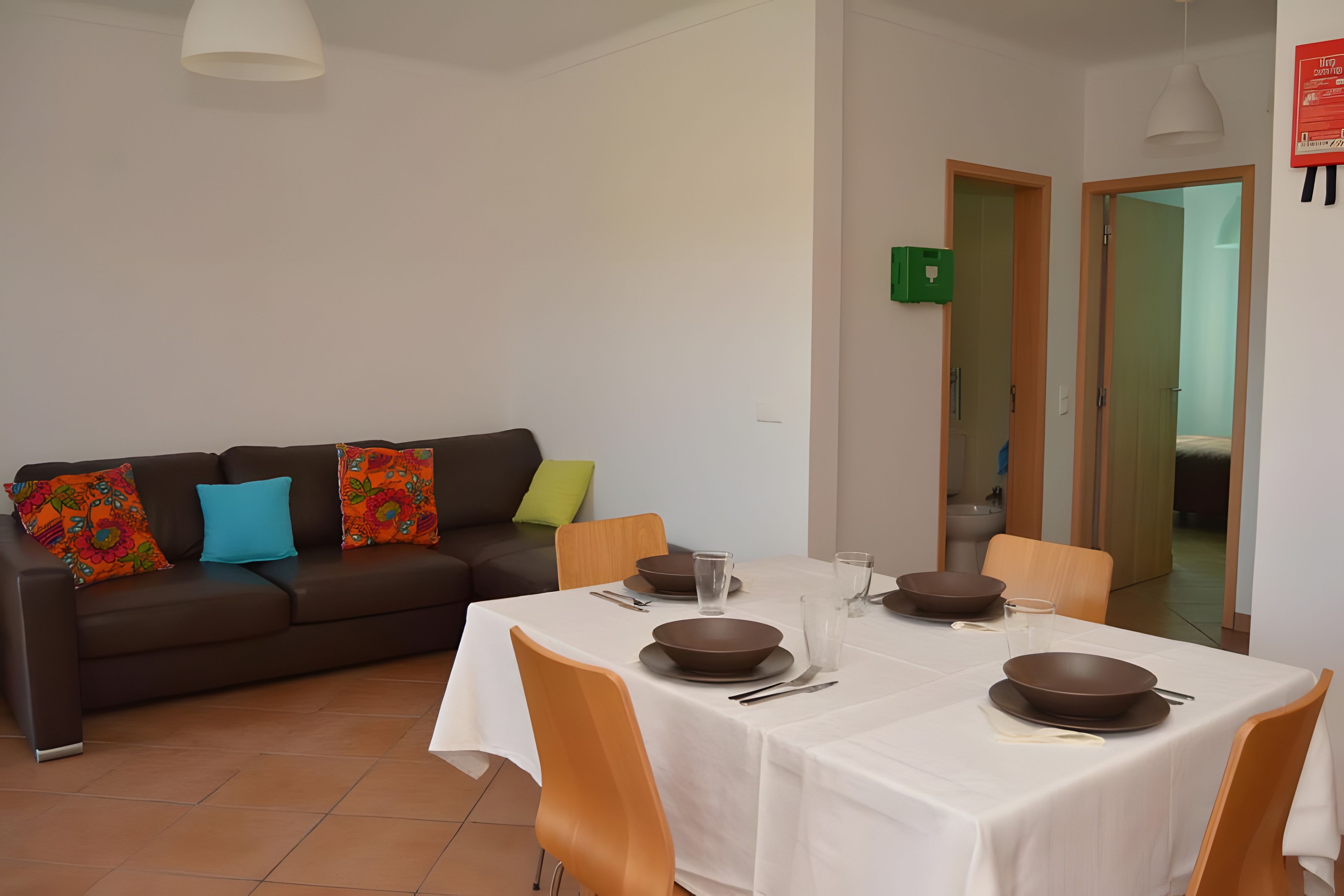 Un coin repas avec une table dressée pour quatre personnes, un canapé agrémenté de coussins colorés et une cuisine visible à travers une porte ouverte.