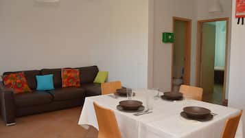 Un coin repas avec une table dressée pour quatre personnes, un canapé agrémenté de coussins colorés et une cuisine visible à travers une porte ouverte.