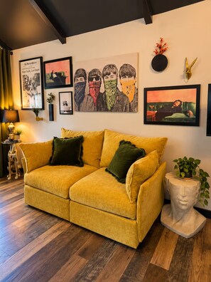 A room with a yellow sofa, a lamp, framed artwork, and a statue. - Eclectic Tiny Home, Private Hot Tub, Shared Pool, In Between Beach & Downtown (Wilmington)