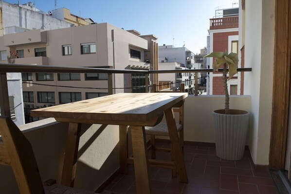 En balkon med et træbord og stole, en potteplante og udsigt til omkringliggende bygninger.