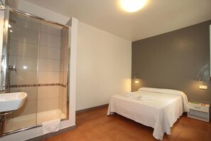 Et hotelværelse med bruser, seng, sengebord og vægmonteret lampe.