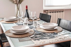 A dining table set with plates, bowls, cutlery, and wine glasses, with a bottle of wine and a vase with dried plants in the background. - New Listing (Roma)