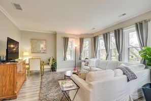 A living room with a white sofa, a wooden cabinet, a glass coffee table, and a large window with curtains.