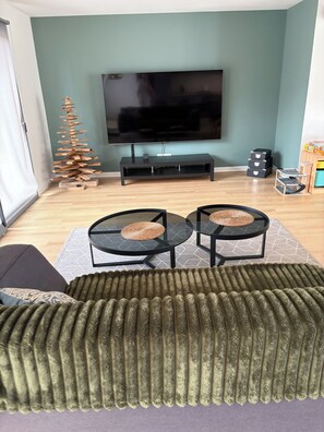 A living room with a flat-screen TV, two round tables with woven stools, a green sofa, and a wooden Christmas tree. - New Listing (Sainte-Hélène)