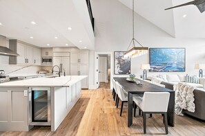 Eine moderne Küche mit Kochinsel, eine Essecke mit dunklem Holztisch und ein Wohnbereich mit Sofa und großem Landschaftsbild.