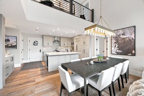 A modern kitchen with a central island, white cabinetry, and a dining area with a dark table and white chairs. - New Remodel Penthouse by InvitedHome | Walk to Ski/Town, Hot Tub, Pool, Gym (Beaver Creek)