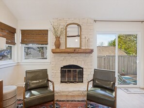 Une pièce avec une cheminée, deux fauteuils en cuir, un miroir et une vue sur l’extérieur, accessible par une porte-fenêtre coulissante.