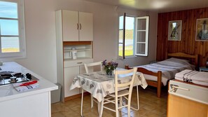 A compact room with a kitchenette, a dining table with chairs, a bed with a wooden headboard, and a window with a view of the beach.