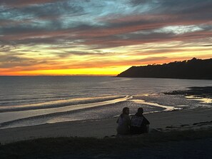 A beach scene with two people sitting on the sand, overlooking the ocean during sunset.