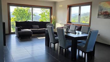 Een moderne woonkamer met een bank, eettafel, stoelen en grote ramen met uitzicht op groen.