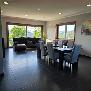 Un salon moderne comprenant un canapé, une table à manger, des chaises et de grandes fenêtres donnant sur un paysage verdoyant.