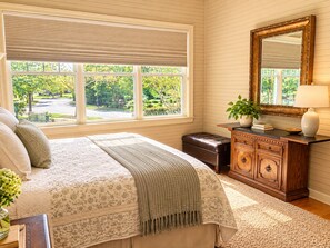 Un dormitorio con un ventanal grande, una cama con cubrecama floral, un aparador de madera y un espejo.