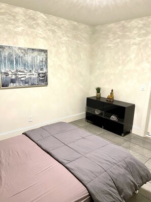 Une chambre à coucher avec un lit, un tableau, un meuble noir pour la télévision sur lequel sont posés divers objets, et une prise électrique au mur.