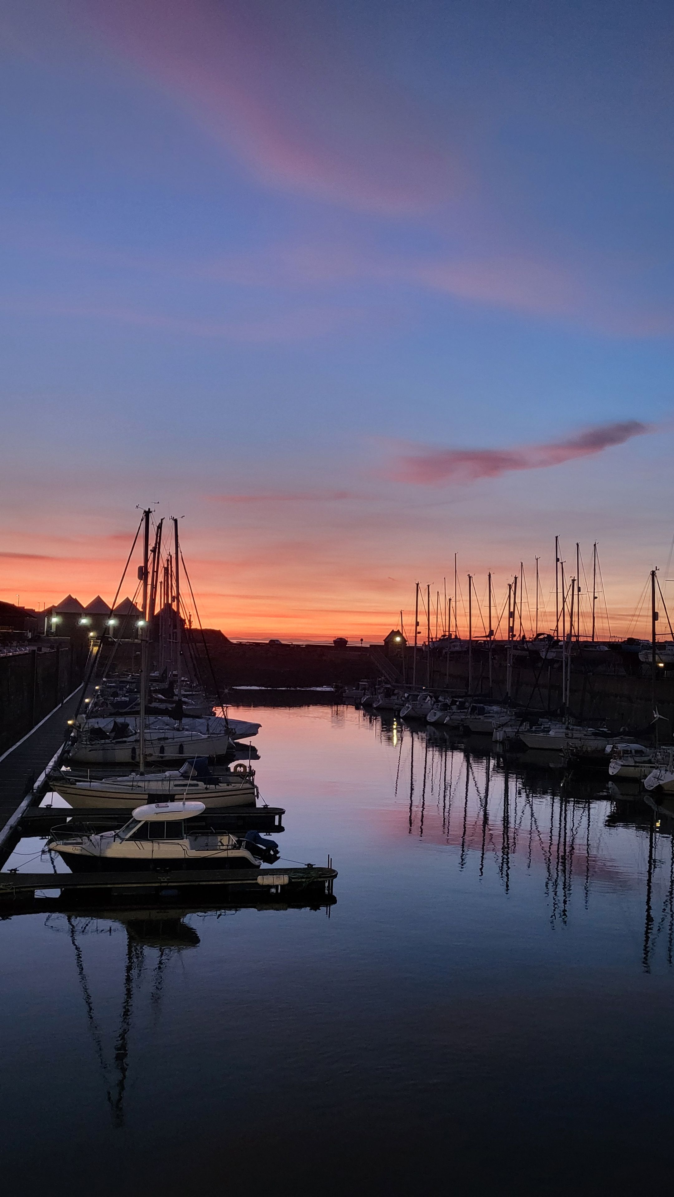 A marina at sunset with boats docked, calm water, and a clear sky.
