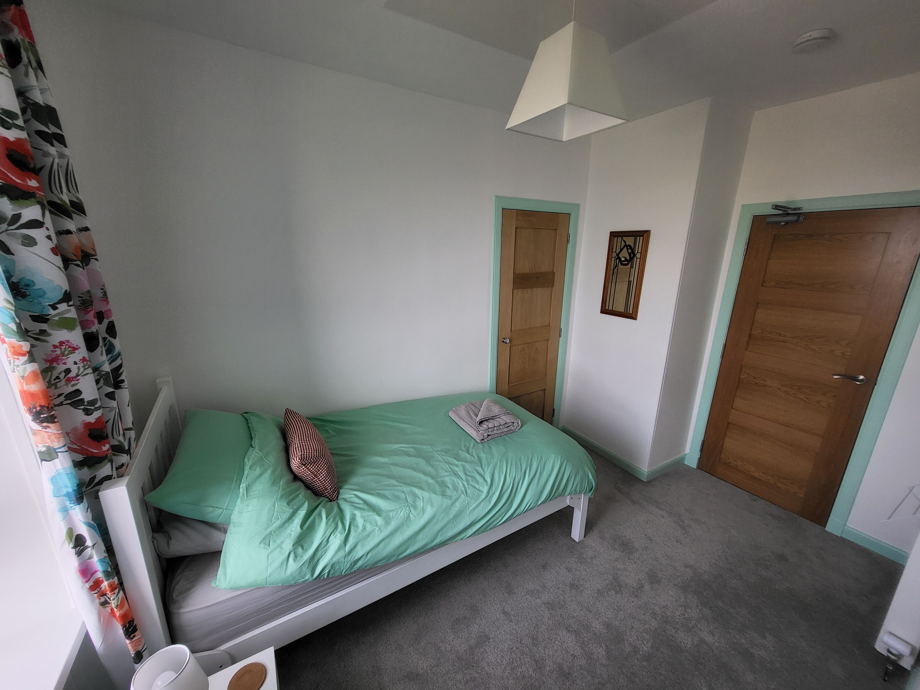A single bed with a green bedspread, a pillow, and a folded towel. There are two doors, one with a mirror above it, and a radiator on the floor.
