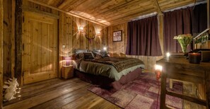 Una habitación de madera con una cama, una mesita de noche, una mesa con lámpara y un aplique de pared.