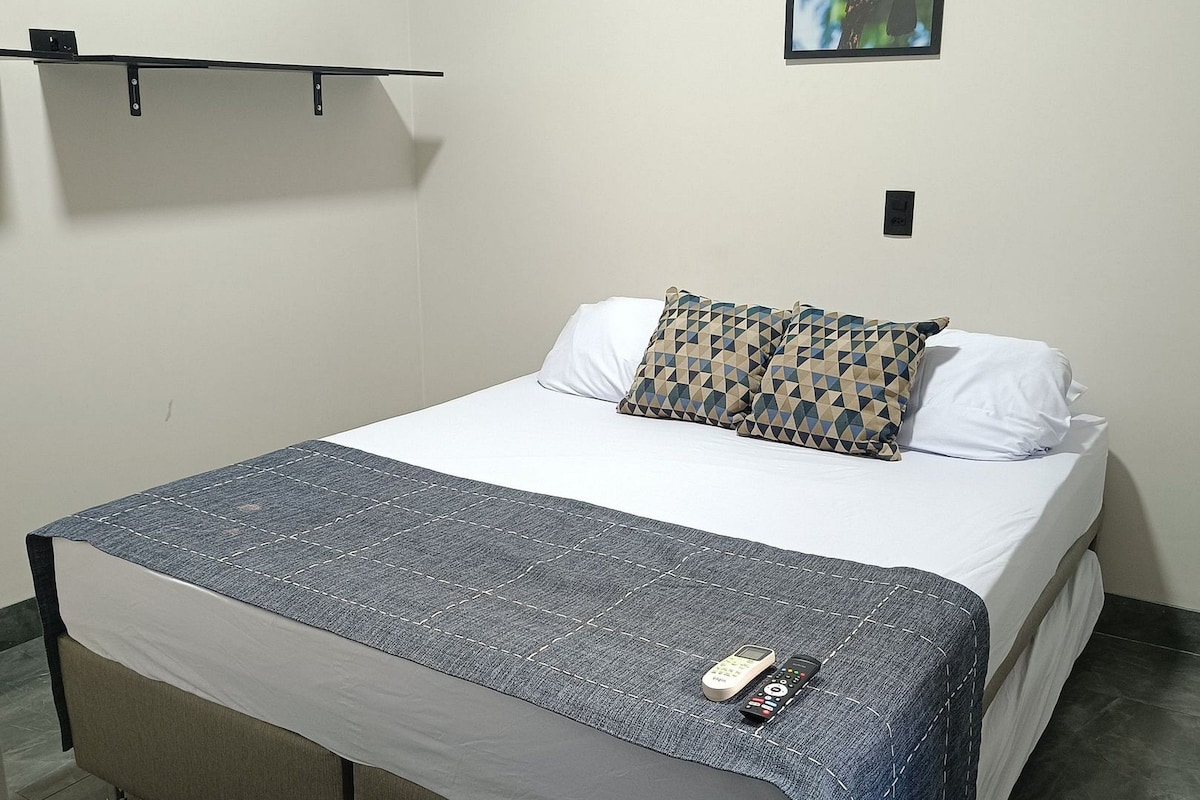 A single bed with a grey mattress and a patterned pillow, a remote control on the bed, and a framed picture of a toucan on the wall.