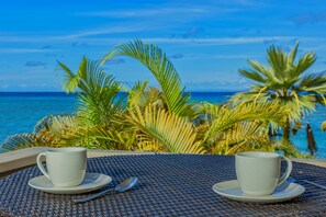 Deux tasses blanches sur une table en fibres tressées, avec vue sur l’océan et les palmiers.