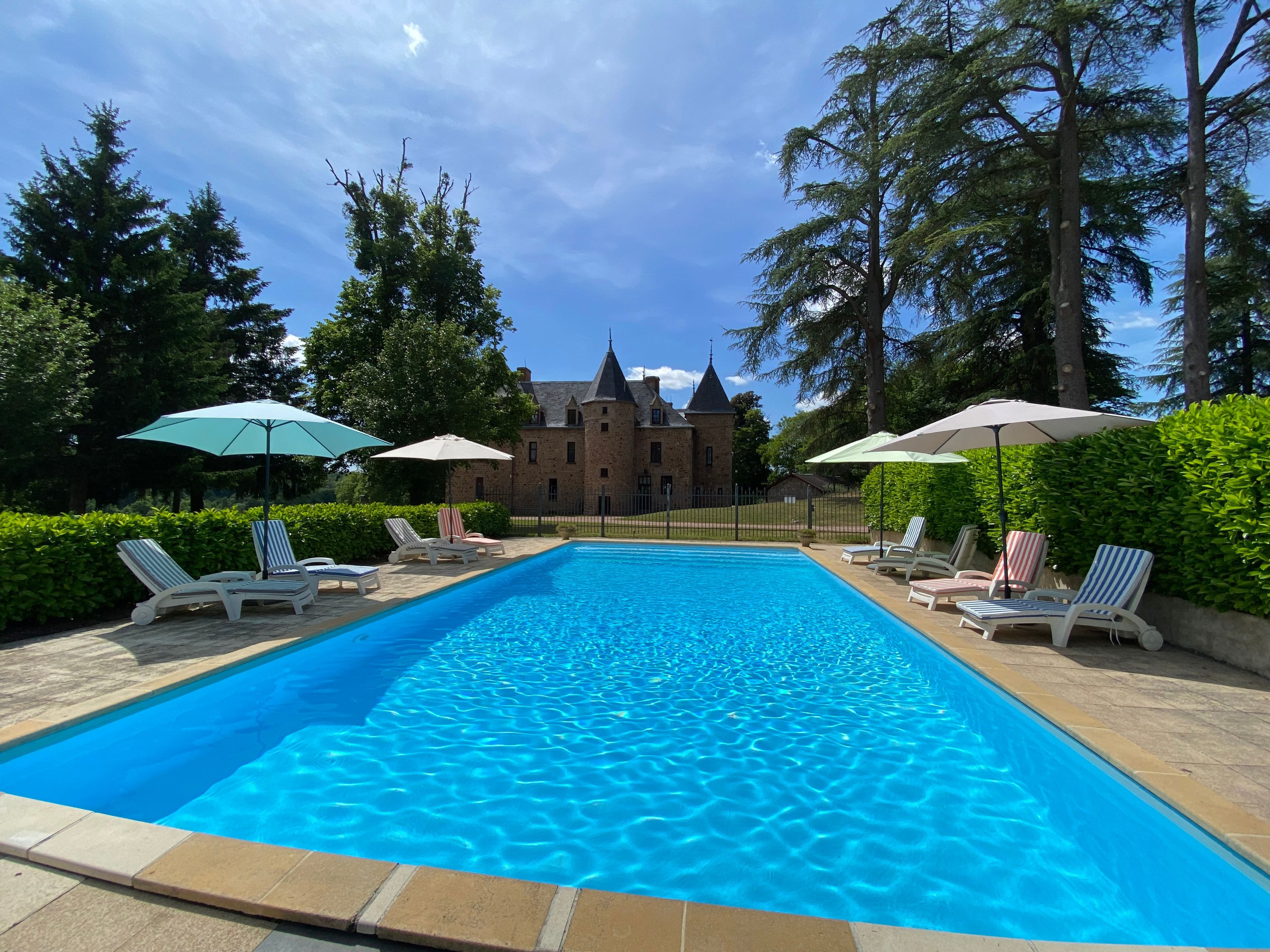 Una piscina rettangolare con lettini e ombrelloni, circondata da siepi e alberi, con un edificio simile a un castello sullo sfondo.
