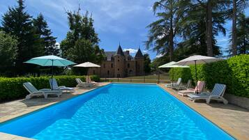 Una piscina rettangolare con lettini e ombrelloni, circondata da siepi e alberi, con un edificio simile a un castello sullo sfondo.