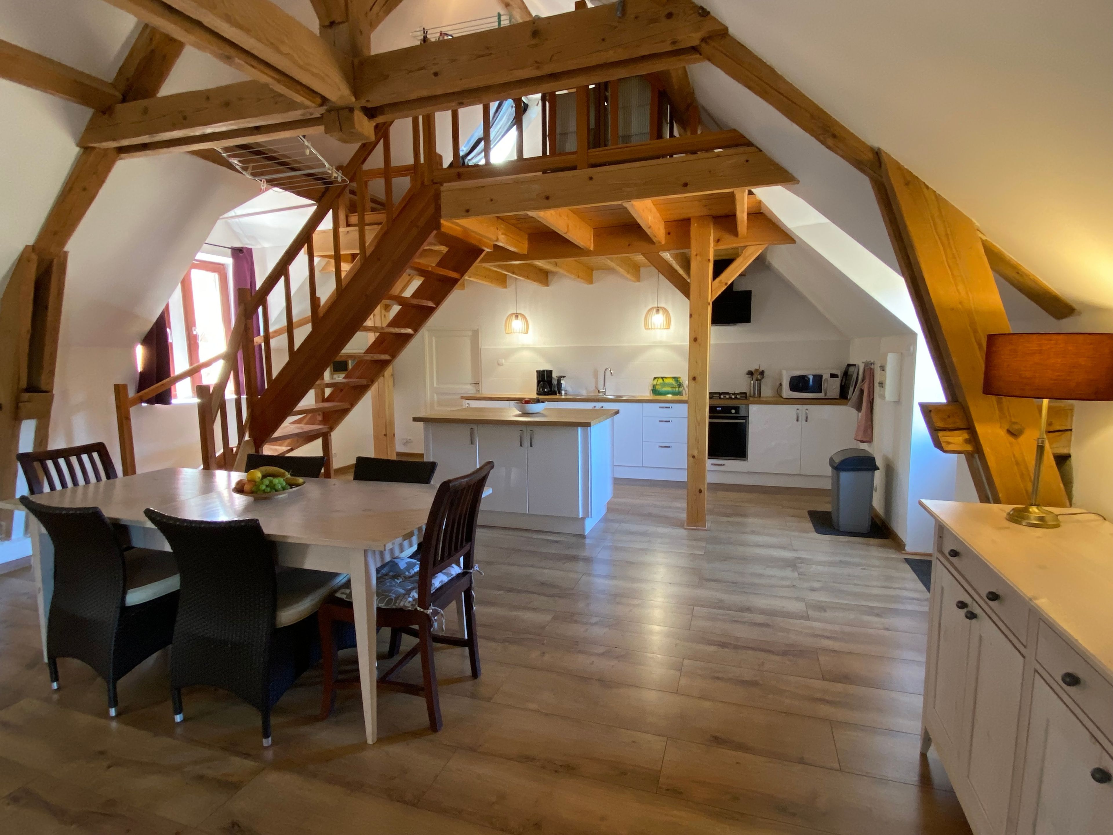 Un loft espacioso con techo alto, vigas de madera a la vista, una escalera central, una zona de comedor con mesa y sillas, y una cocina con armarios blancos y electrodomésticos modernos.