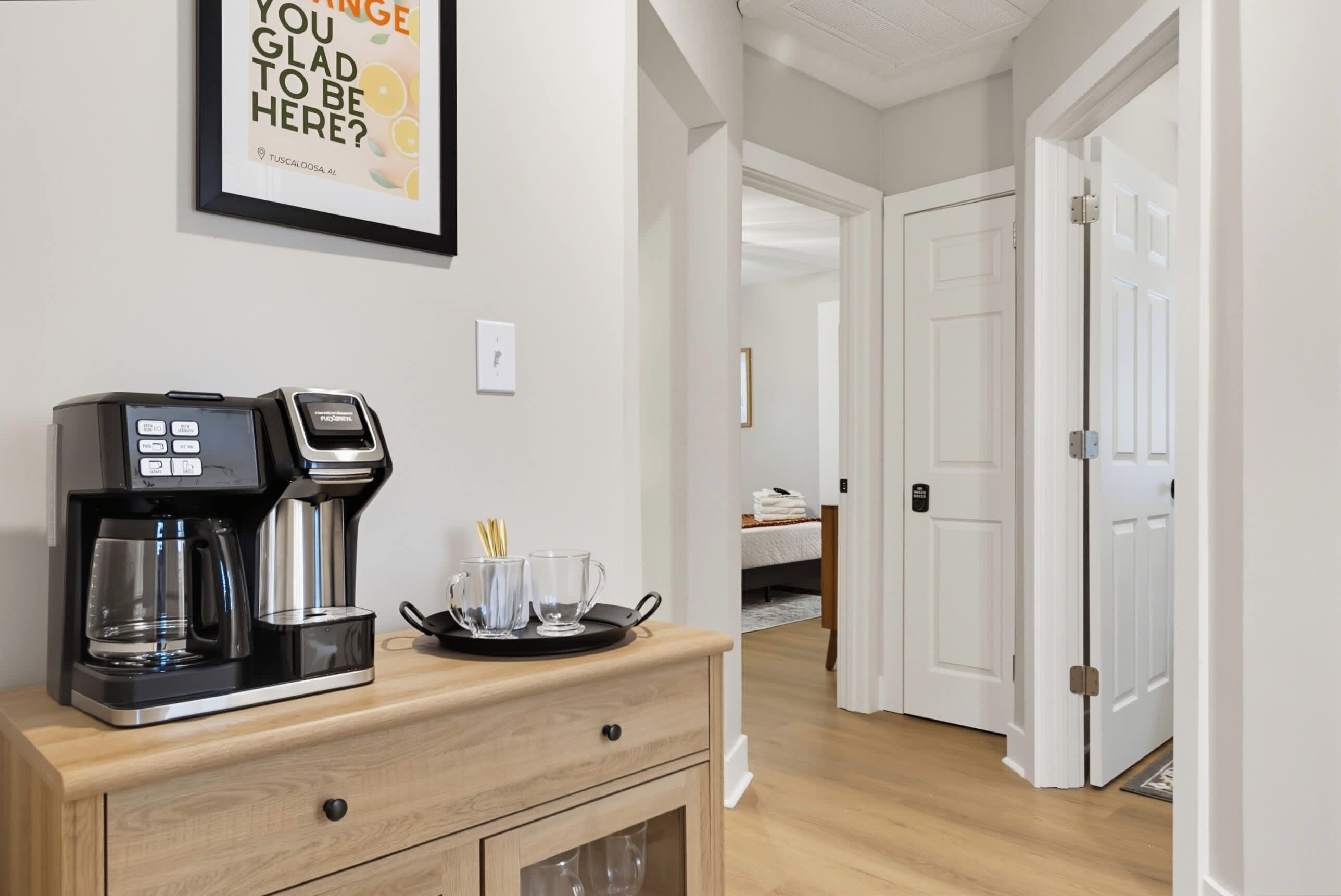 Une chambre d’hôtel avec une machine à café posée sur un comptoir en bois, un panneau encadré accroché au mur et une vue sur la chambre adjacente.