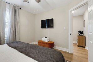 Quarto de hotel com cama, televisão fixada na parede, um banquinho de madeira e uma máquina de café.