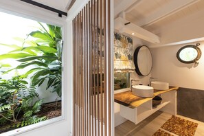 Een moderne badkamer met een houten wastafelmeubel, twee wastafels, een grote spiegel en een raam met tropische planten.