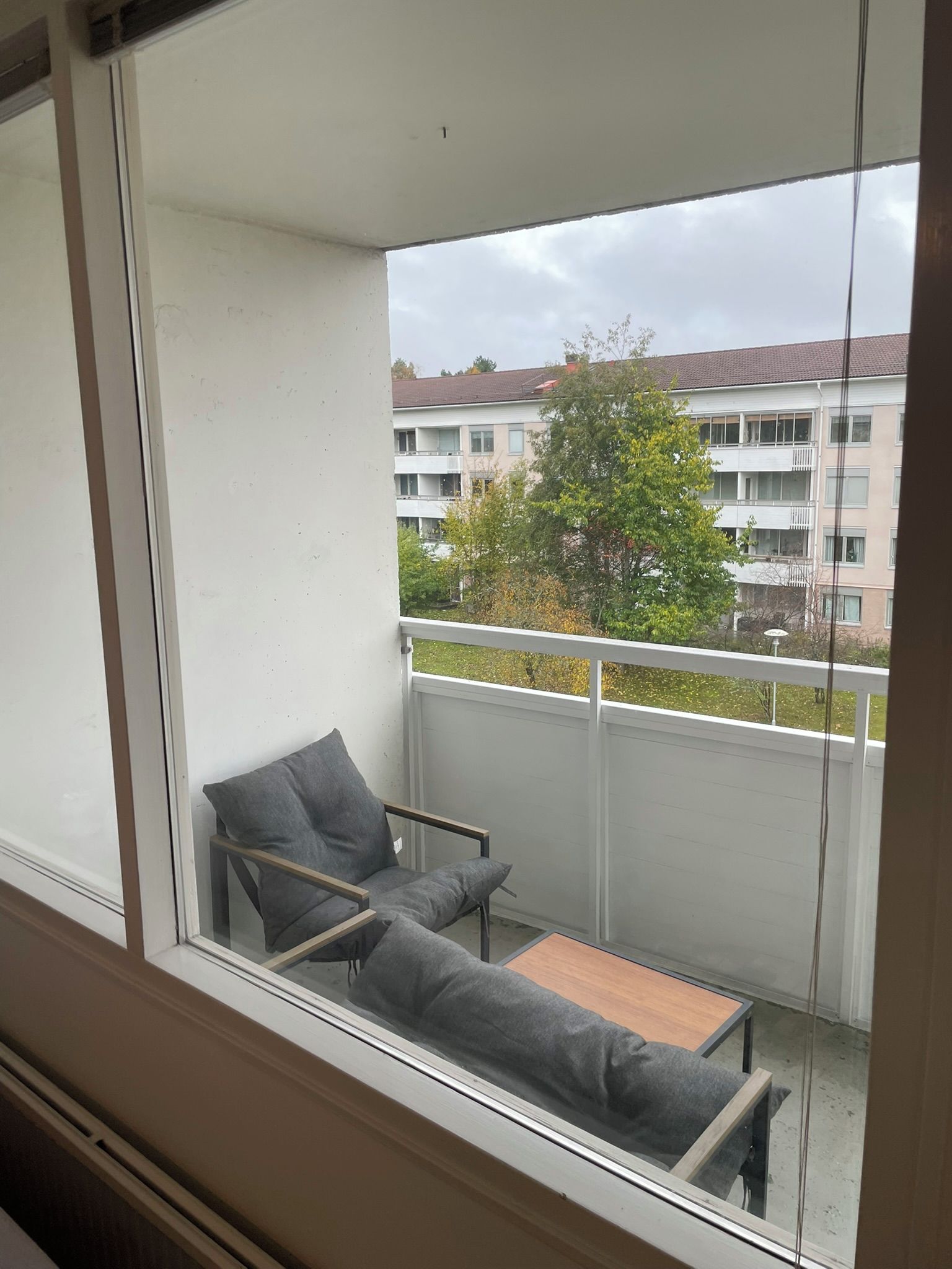 Un balcón con un sofá gris y una mesita, con vistas a una zona residencial con edificios y árboles.