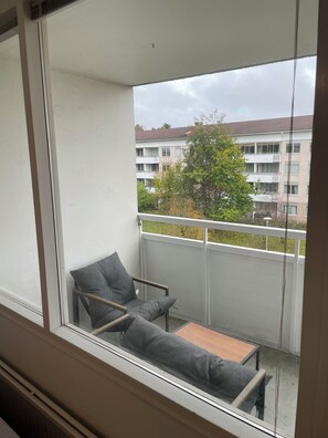 A balcony with a grey sofa and a small table, overlooking a residential area with buildings and trees. - New Listing (Handen)