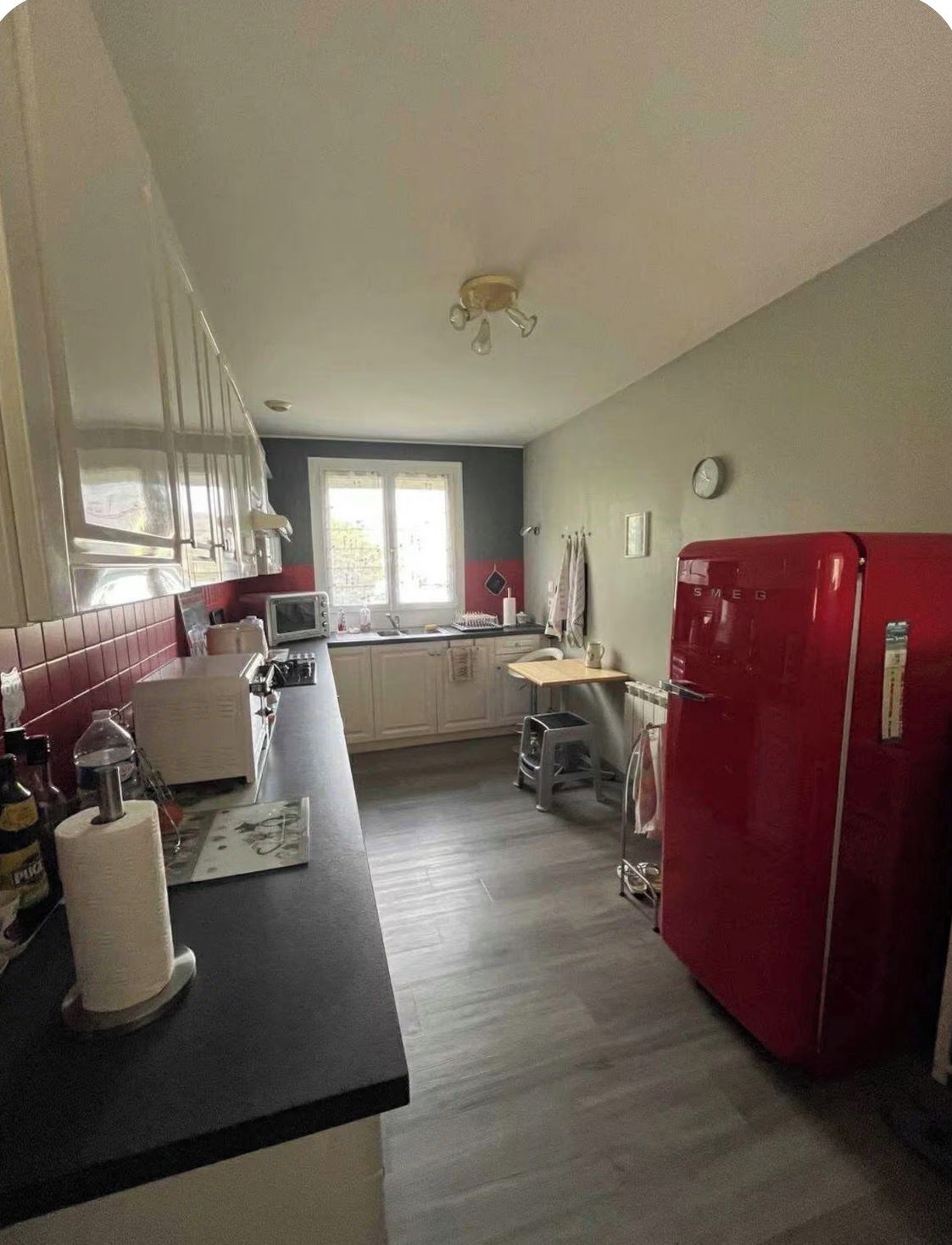 A kitchen with a red refrigerator, white cabinets, a window, and a clock on the wall.