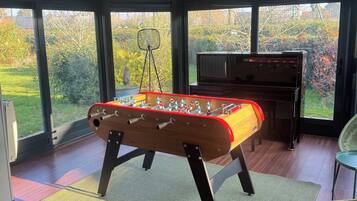A room with a foosball table, a piano, and large windows offering a view of the outdoors.