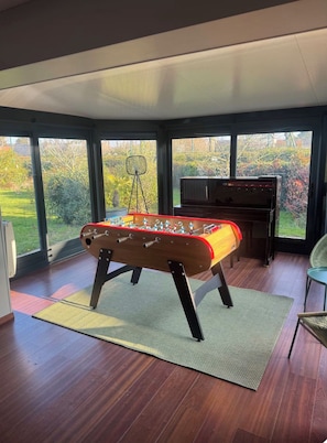 A room with a foosball table, a piano, and large windows offering a view of the outdoors.