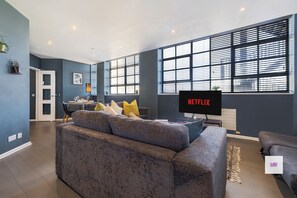 Ein modernes Wohnzimmer mit einem grauen Sofa, einem Flachbildfernseher und großen Fenstern.