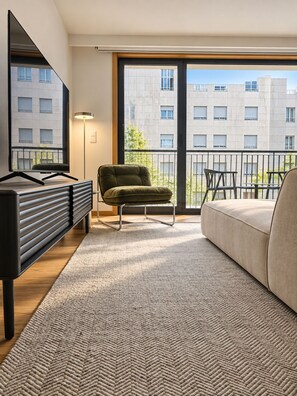 Un salon moderne avec un canapé, un fauteuil vert, un meuble pour la télévision et une grande fenêtre donnant sur des immeubles.