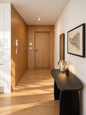 Un couloir avec des portes en bois, une œuvre d’art représentant un paysage urbain encadrée et une table avec une lampe.