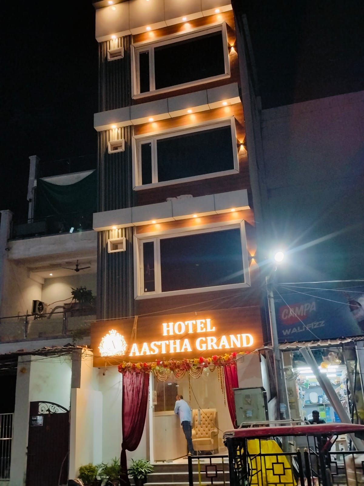 Un hotel llamado 'Hotel Aastha Grand' con un letrero, un balcón con una silla y un escaparate con un anuncio para 'Campa WalfiZZ'.