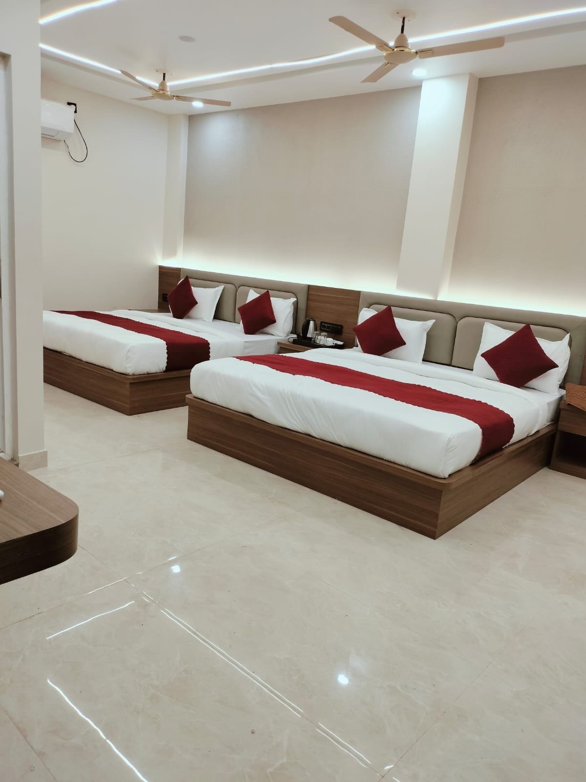 Habitación de hotel con dos camas, cada una con cabecero de madera, ropa blanca y almohadas rojas. Cuenta con ventilador de techo, mesita de noche con lámpara y aire acondicionado instalado en la pared.