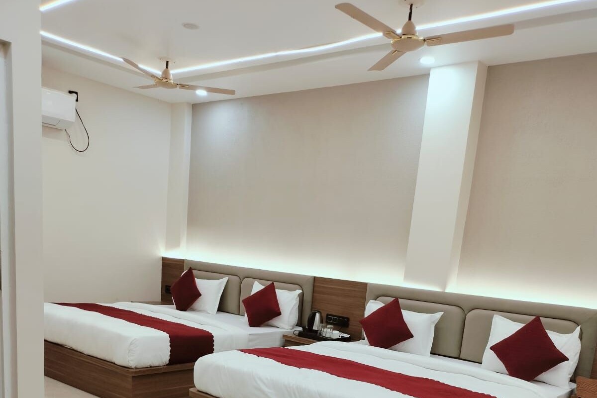 A hotel room with two beds, each with a wooden headboard, white bedding, and red pillows. There is a ceiling fan, a bedside table with a lamp, and a wall-mounted air conditioning unit.