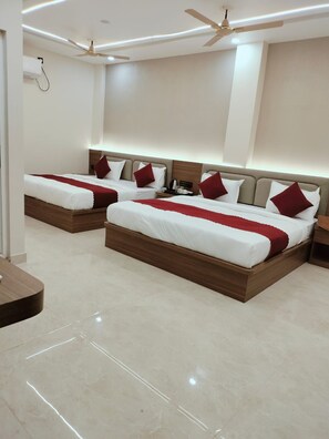A hotel room with two beds, each with a wooden headboard, white bedding, and red pillows. There is a ceiling fan, a bedside table with a lamp, and a wall-mounted air conditioning unit. - Hotel Aastha Grand (Varanasi)