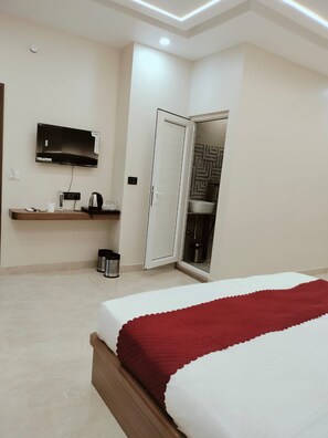 A hotel room with a bed, a TV mounted on the wall, a small table with a kettle, and a door leading to a bathroom. - Hotel Aastha Grand (Varanasi)
